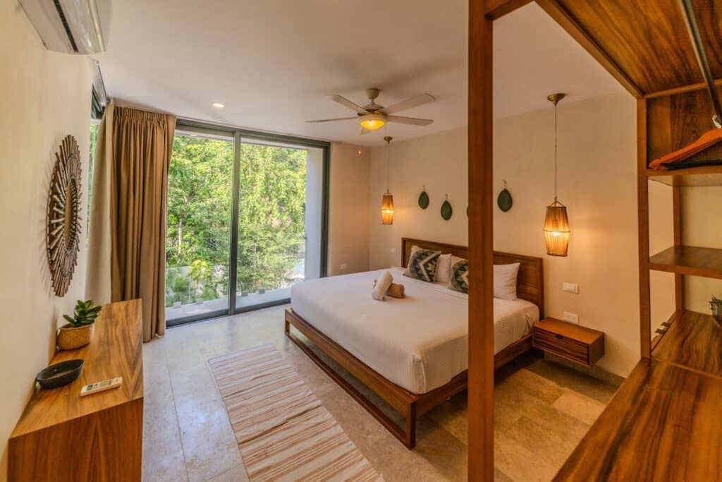 Bedroom with large glass sliding door, wooden furniture, pendant lights, and tropical greenery visible outside at Villa Las Almas in Aldea Zama, Tulum.