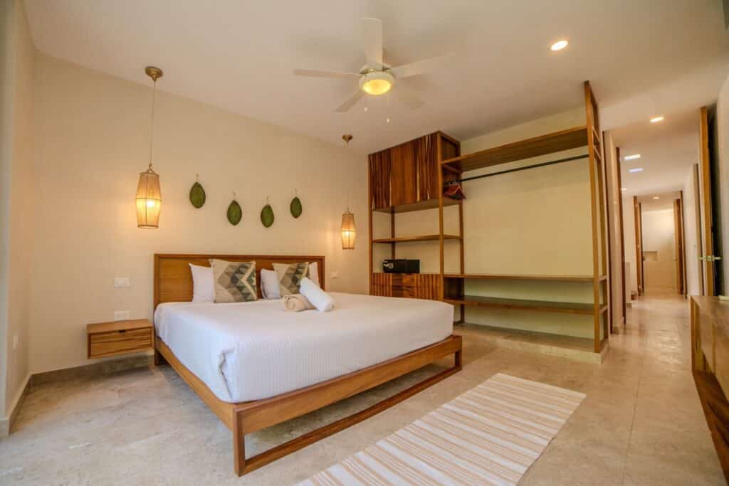 Bedroom with a wooden bed frame, white linens, pendant lighting, and a built-in open closet at Villa Las Almas in Aldea Zama, Tulum.