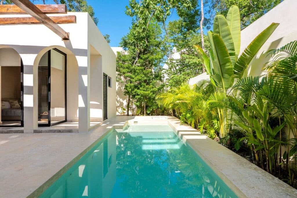View of a narrow rectangular swimming pool with tropical plants on the side and a modern white building at Villa Serenia in Kaybe Tulum.
