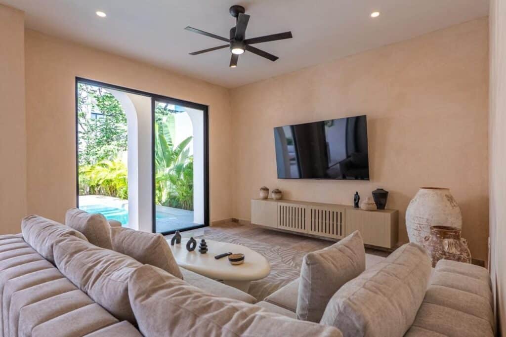Living room with beige walls, a large sectional sofa, a wall-mounted TV, and a sliding glass door leading to a garden with pool at Villa Serenia in Kaybe Tulum.