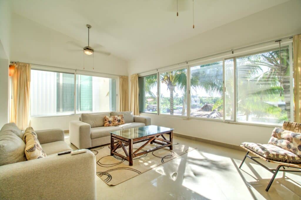 Living room with large windows showing palm trees and sunlight, furnished with beige sofas, a wooden coffee table, and curtains at property name in area.