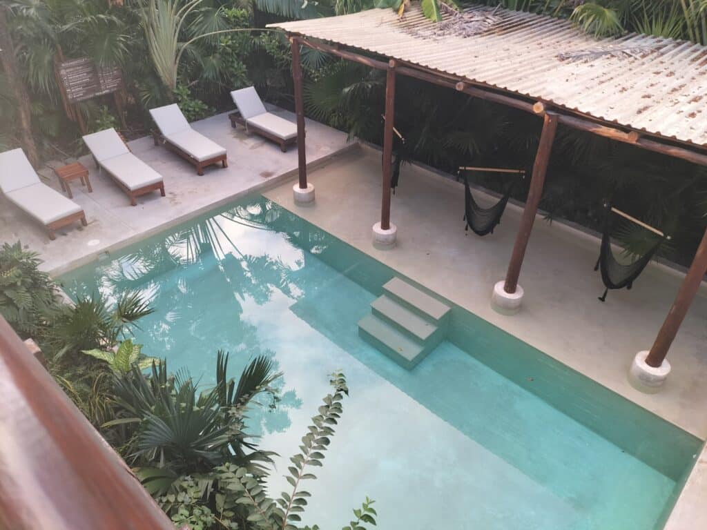 View of a small outdoor pool with steps, surrounded by a concrete deck, white lounge chairs, and lush tropical greenery in Tulum Centro.