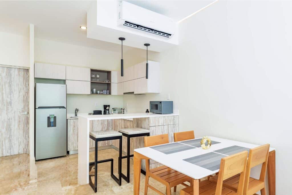 Modern kitchen with white cabinetry, stainless steel appliances, and a dining area with wooden chairs and a white table at The Elle in Aldea Zama, Tulum.