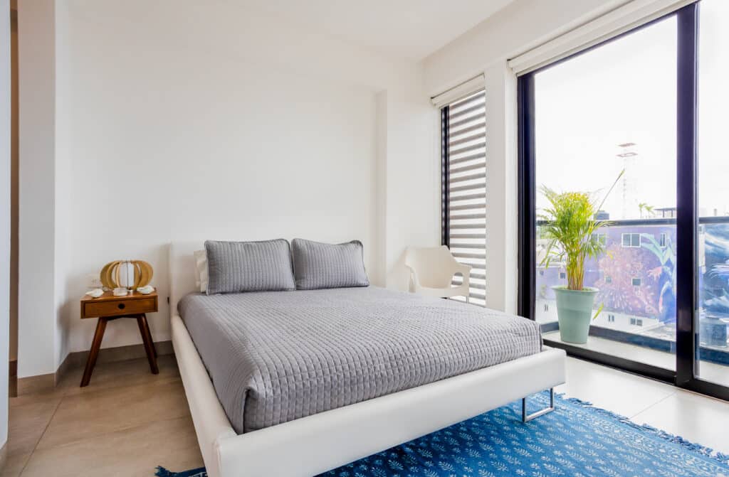 Bedroom with a large window, gray bedding, and wooden nightstand in a modern apartment at Playa del Carmen at New Icono B407 3-Bedroom Condo.
