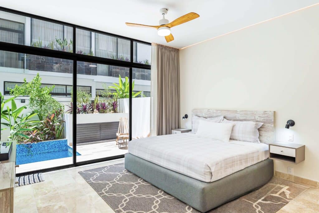 Modern bedroom with large glass doors opening to a balcony with lush plants and a pool in Tulum at The Elle.