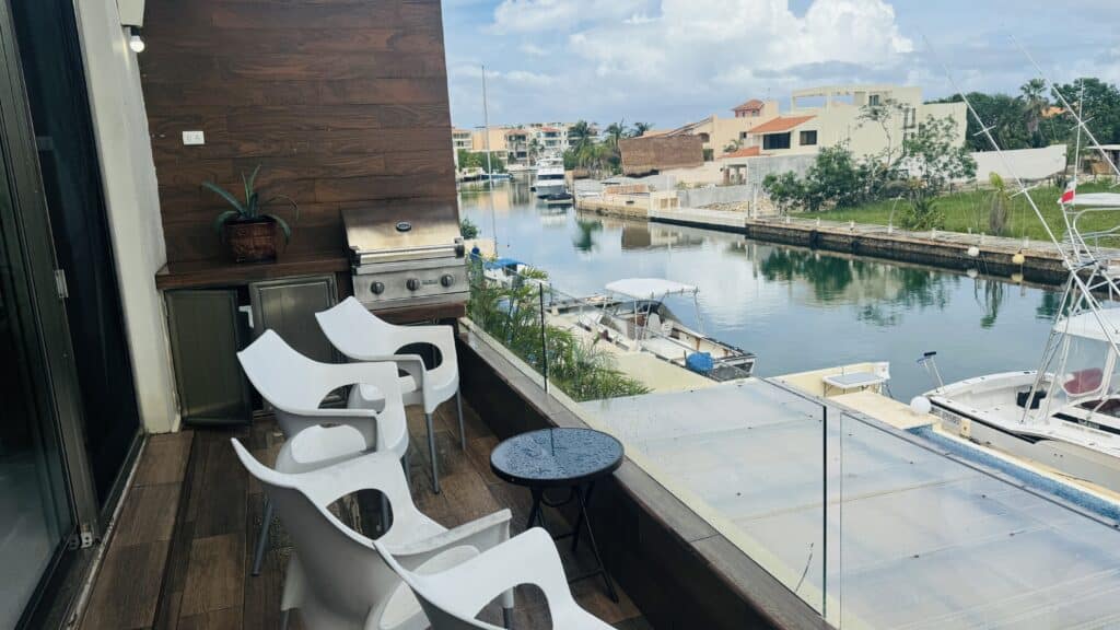 Balcony with white chairs and black glass table overlooking the canal and boats, with neighboring houses and greenery in Puerto Aventuras at Regatta 4 4-Bedroom Penthouse.