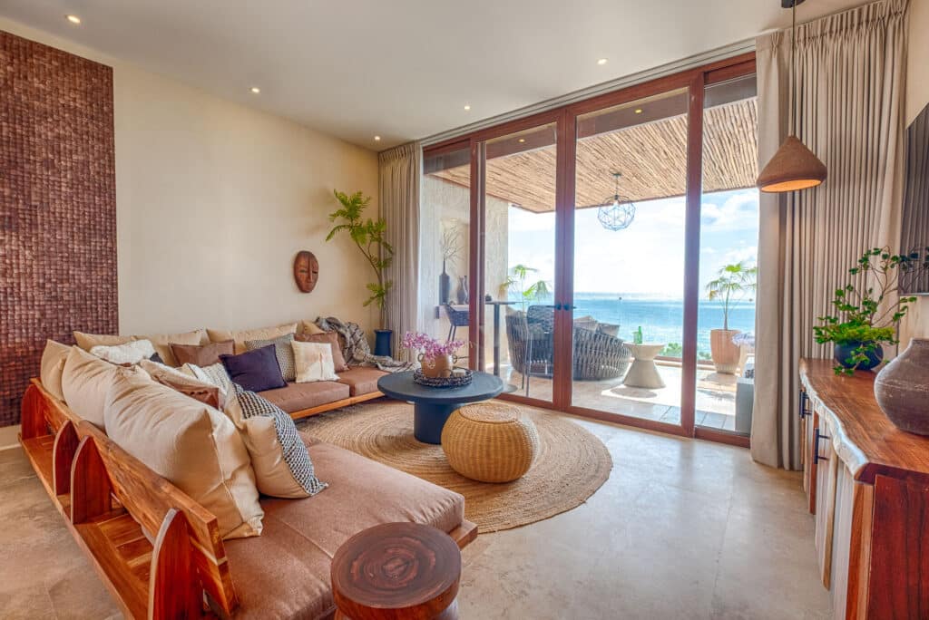 Living room with a beige sectional sofa, wooden accents, and large glass doors opening to a balcony with ocean views in Tulum at Kaoba.
