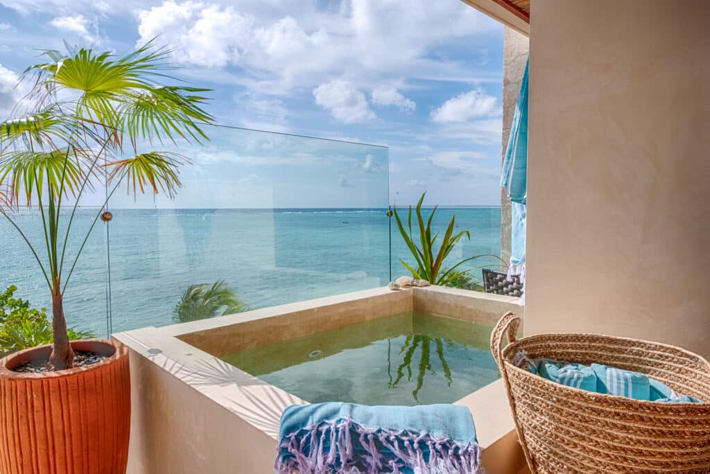 View of the ocean from a balcony with potted plants and wicker furniture, overlooking a glass railing and blue sky at Kaoba in Tankah Bay, Tulum.