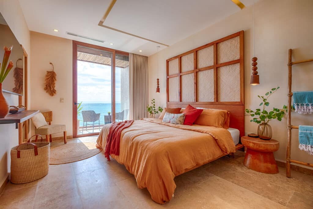 Bedroom with a large glass door leading to a balcony overlooking the ocean, decorated with earth-tone bedding, plants, and woven furniture at Kaoba in Tankah Bay, Tulum.
