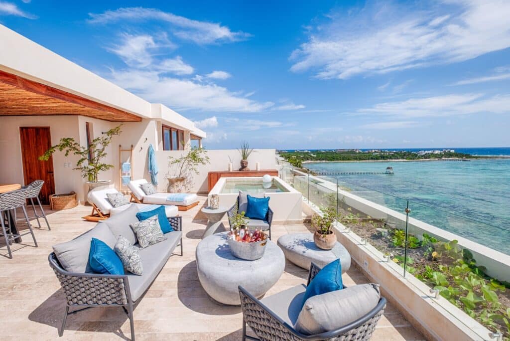 Rooftop terrace with outdoor seating, umbrellas, greenery, and a view of the ocean and pier on a sunny day in Tulum at Kaoba.