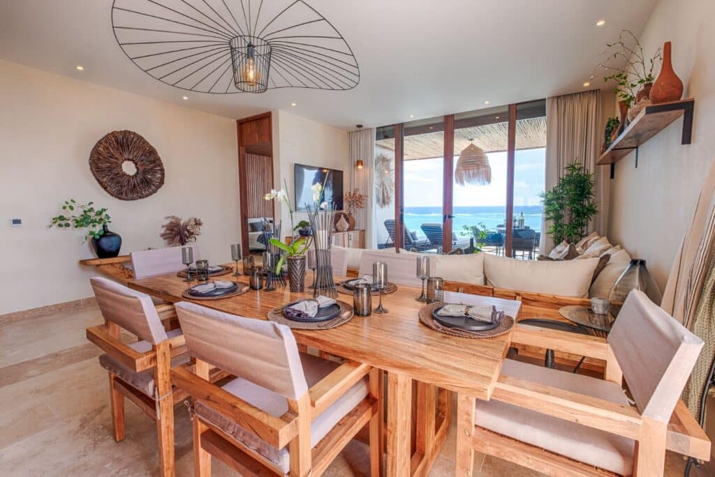 Interior view of a dining area with a wooden table, beige chairs, and a large glass door overlooking a beach and ocean in Tulum at Kaoba.