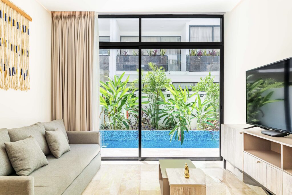 Living room with a large glass sliding door, beige sofa, and tropical plants outside in Tulum at The Elle.
