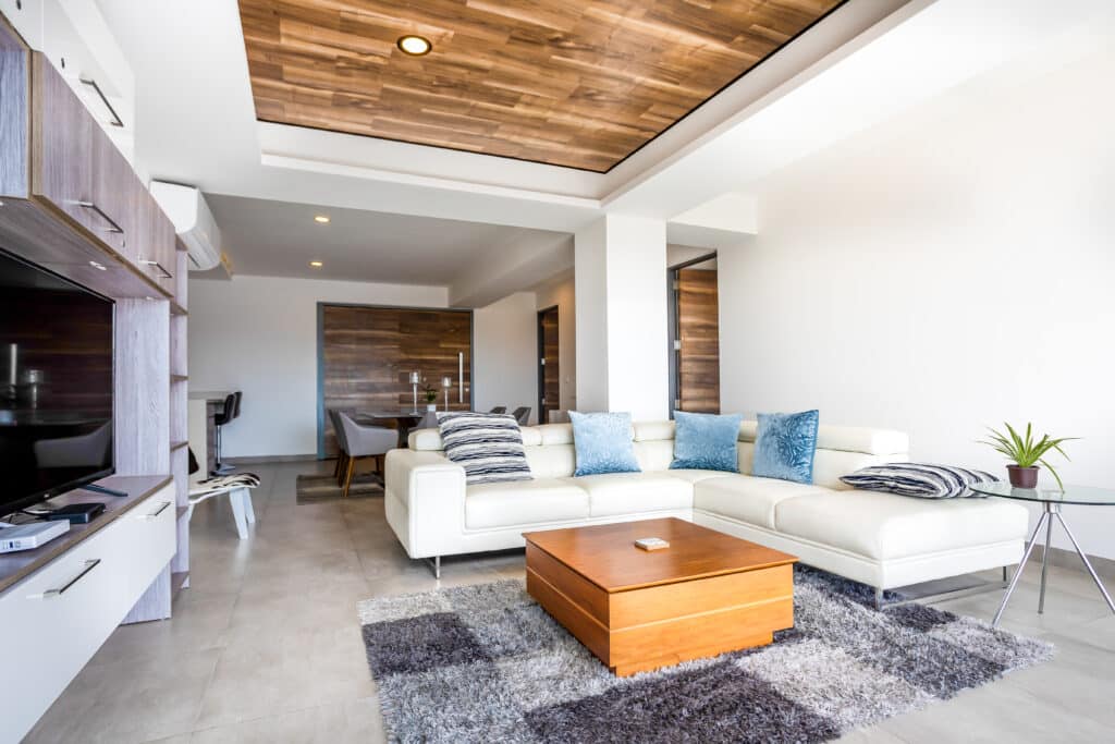 Living room with white sectional sofa, wooden coffee table, TV unit, and accent pillows in a modern condo with wooden and white ceiling accents in Playa del Carmen.
