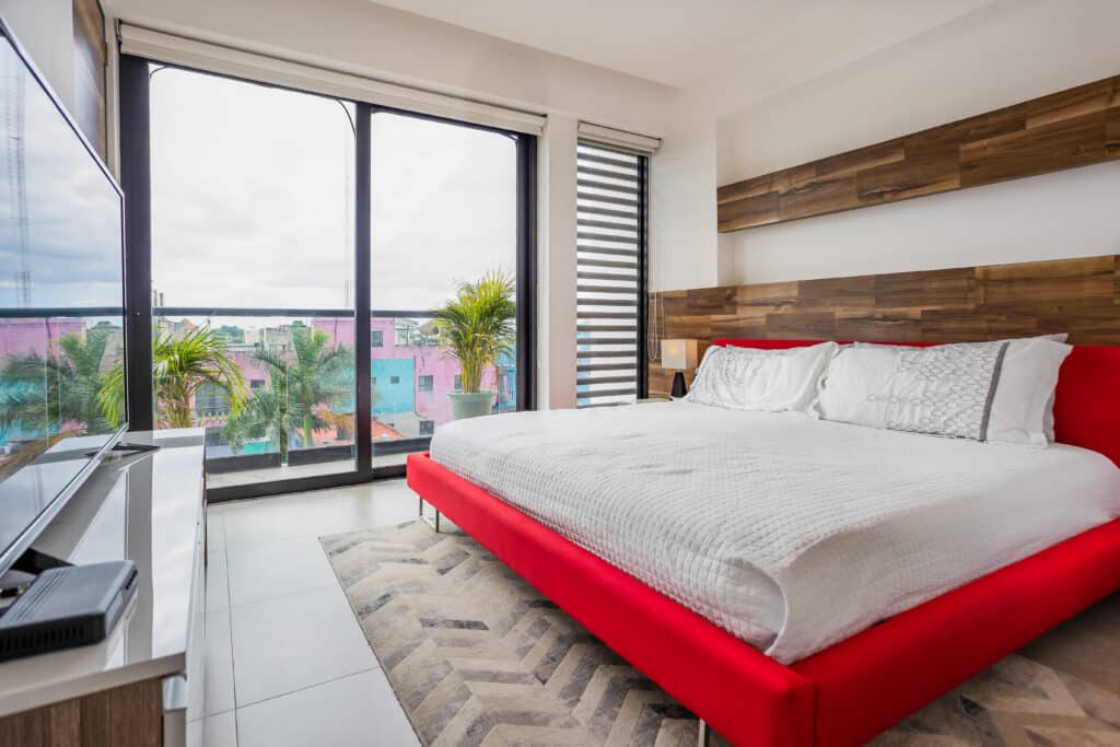 Bedroom with large glass sliding doors opening to a balcony, view of colorful buildings and trees outside, wooden feature wall, white bedding in Playa del Carmen.