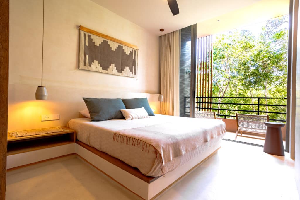 Bedroom with large glass door opening to a balcony with trees, beige walls, ceiling fan, and minimalist furniture in Tulum.
