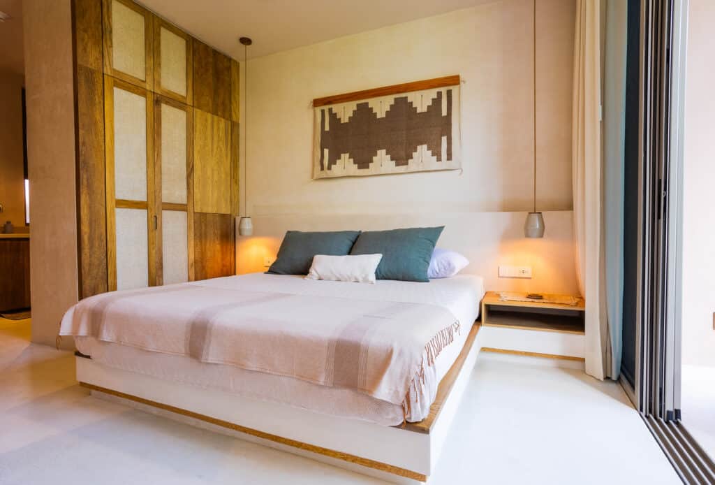 Bedroom with a wooden-paneled wall, hanging textile art, and sliding glass door leading to a balcony at Aflora in Aldea Zama, Tulum.