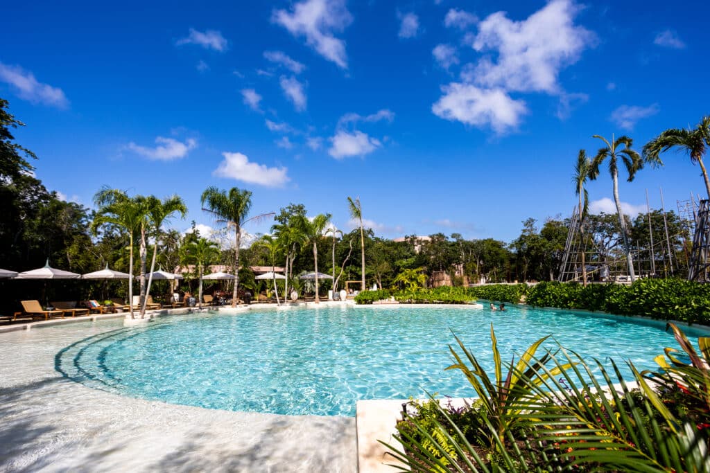 Swimming pool with clear blue water, surrounded by lounge chairs and lush tropical trees, under a bright blue sky in Tulum at Aflora.