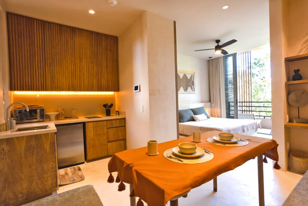 Open-plan kitchen and dining area with wooden cabinets, orange tablecloth, and view of bedroom with balcony and greenery in Tulum at Aflora.