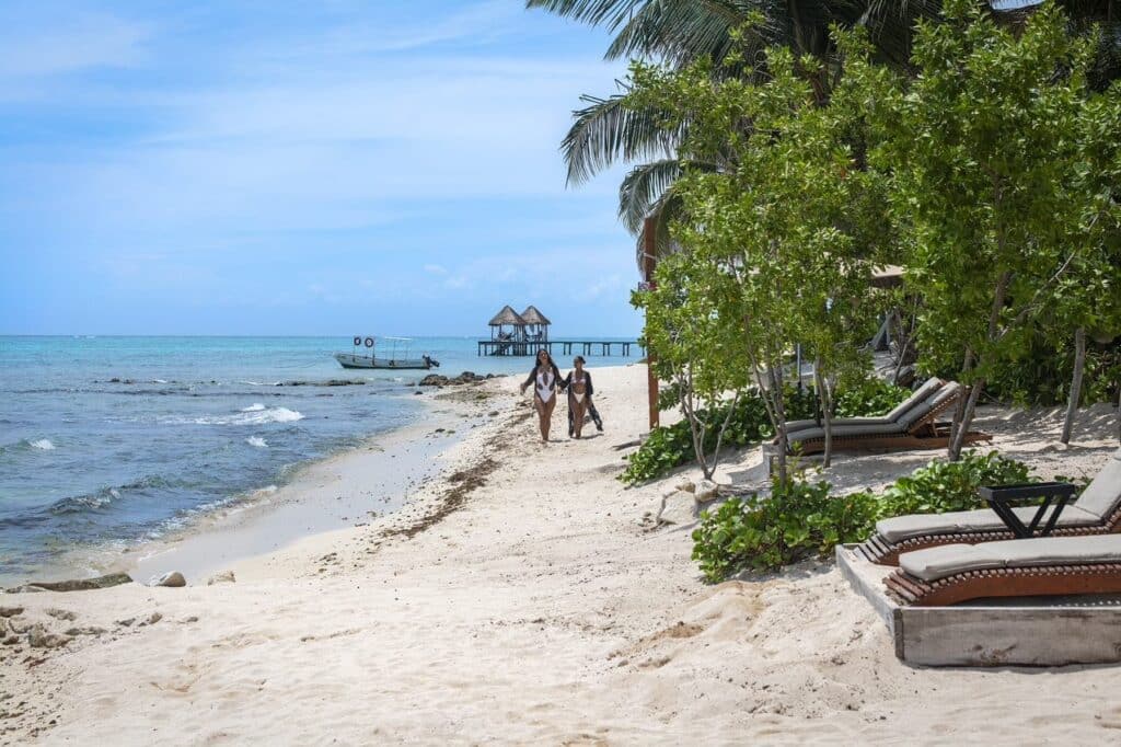 Two women walk along a sandy beach with lounge chairs and lush greenery, overlooking the ocean in Tulum at 21 Room Hotel.