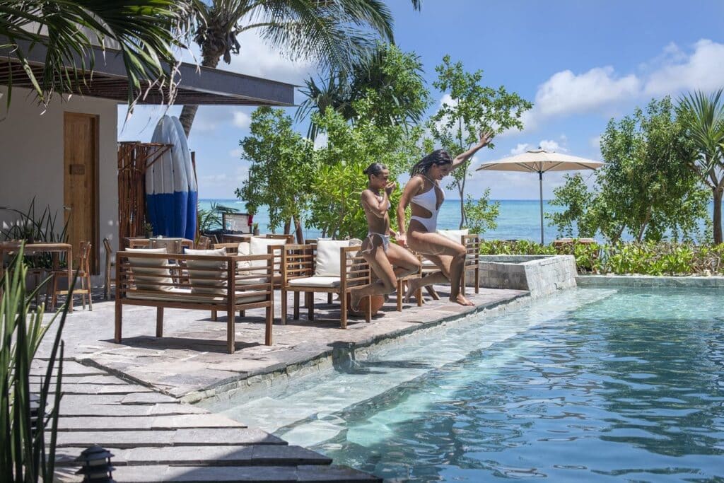 Two women are relaxing by an outdoor swimming pool with ocean views in Tulum, surrounded by trees and sun umbrellas at 21 Room Hotel.