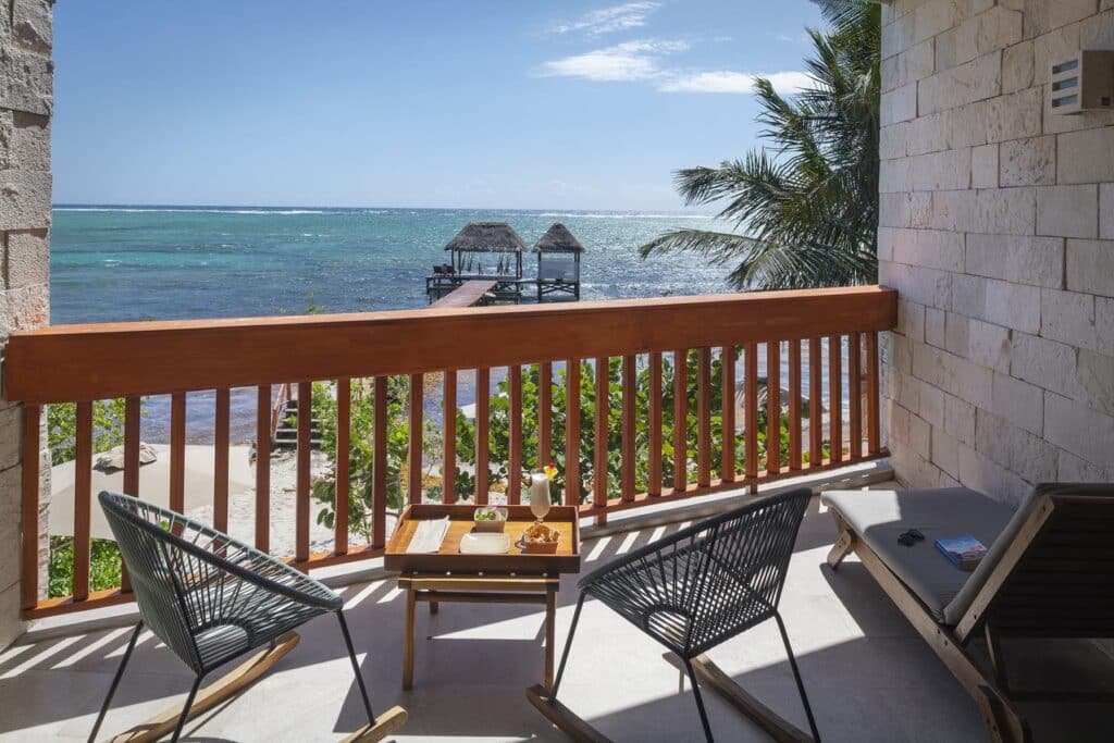 Balcony overlooking the ocean with two lounge chairs, small table with drinks, and a sandy beach with thatched-roof cabanas in Tulum at 21 Room Hotel.