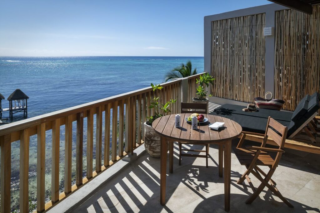 Balcony with wooden railing overlooking the ocean, with outdoor furniture including a round table, chairs, potted plants, and sunshade panels in Tulum at 21 Room Hotel.