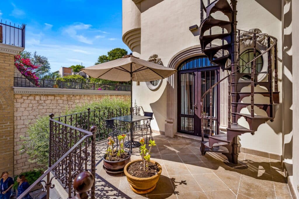 View of a balcony with potted plants, a patio table with an umbrella, a curved stairway, and ornamental architectural details at Casa 28 De Abril in San Miguel de Allende.