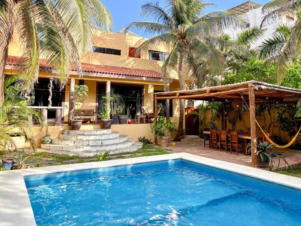 View of a yellow house with a tiled roof, surrounded by palm trees and tropical plants, featuring a swimming pool and outdoor seating area at Casa Tankah Villa in Tulum.