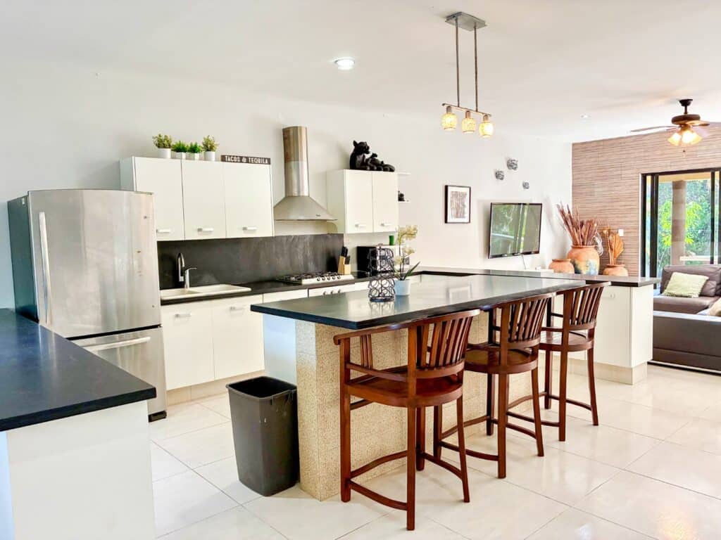 Modern kitchen with white cabinets, black countertops, and stainless steel appliances, open to a living area with a mounted TV in a property near Tankah Bay at Casa Tankah Villa.