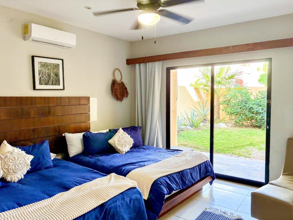 Bedroom with dressed in blue bedding, decorative pillows, sliding glass door opening to a garden in Casa Tankah Villa, Tankah Bay, Tulum.