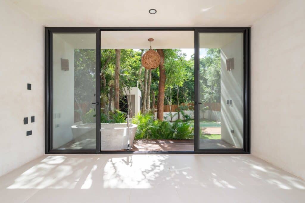 Interior view of a room with a large glass sliding door opening to a lush outdoor garden with trees and plants at Zapote in Tulum.