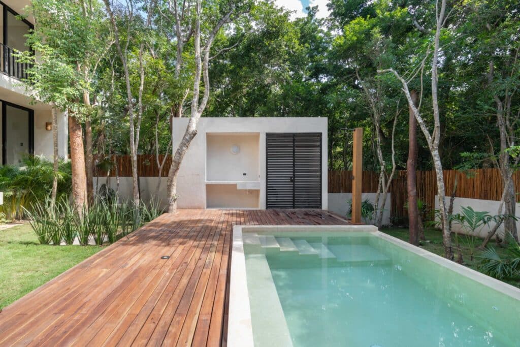 Modern backyard with a small swimming pool, wooden deck, surrounded by lush green trees and a white minimalistic structure in a tropical setting at Zapote in Tulum.