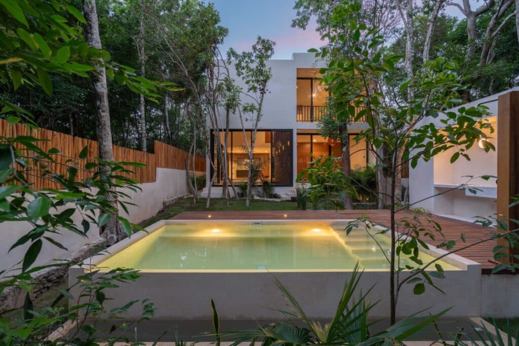 Modern two-story house with large glass windows, surrounded by trees, and a small illuminated swimming pool in the backyard at Zapote in Tulum.