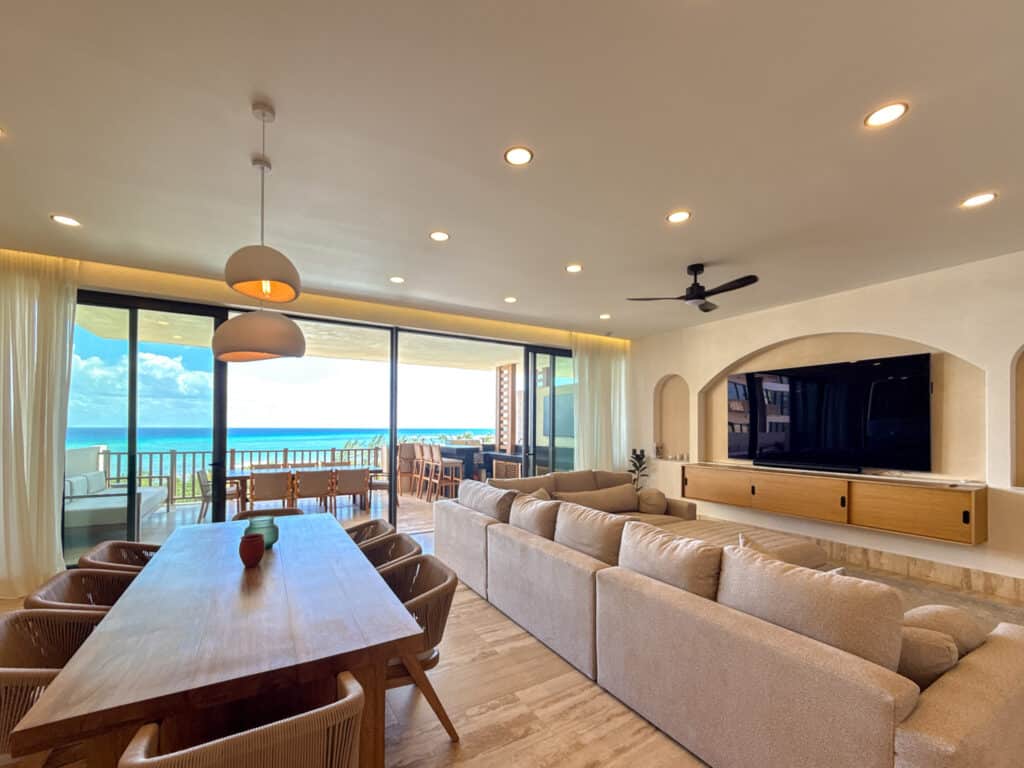 Interior living space with large windows offering ocean views, a beige sectional sofa, wooden dining table, modern pendant lights, and ceiling fan in Corasol, Playa del Carmen.