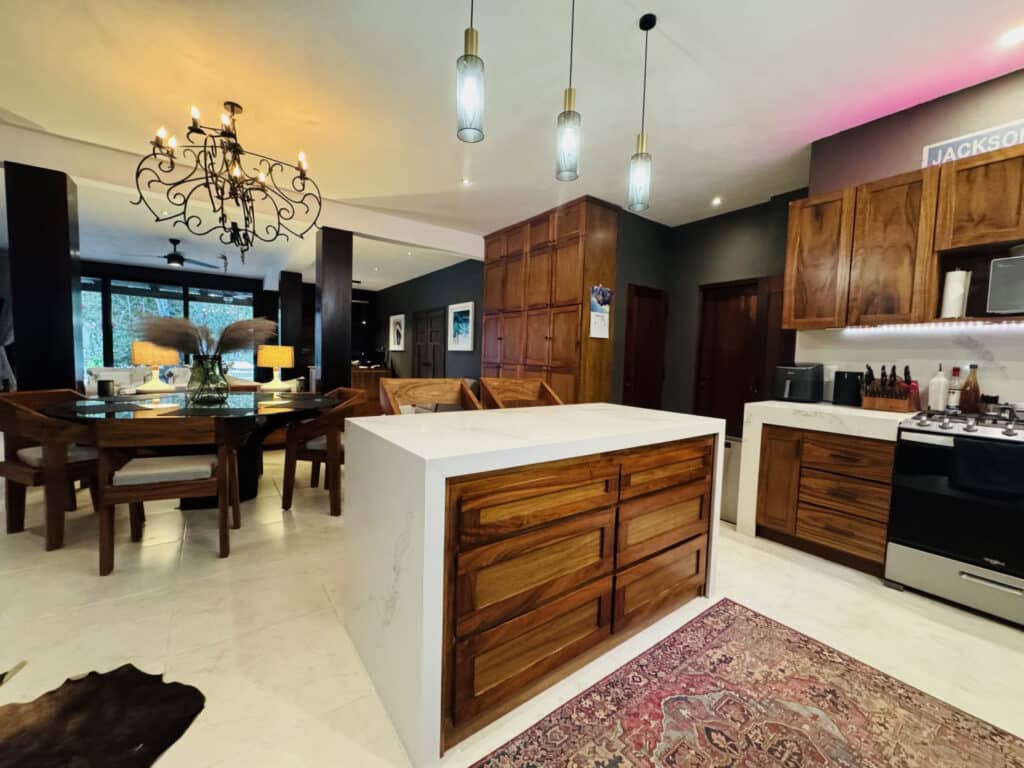 Interior view of a modern kitchen with wooden cabinets, white countertops, pendant lighting, and an open dining area with a chandelier in a home at Hacienda MVO in Akumal.