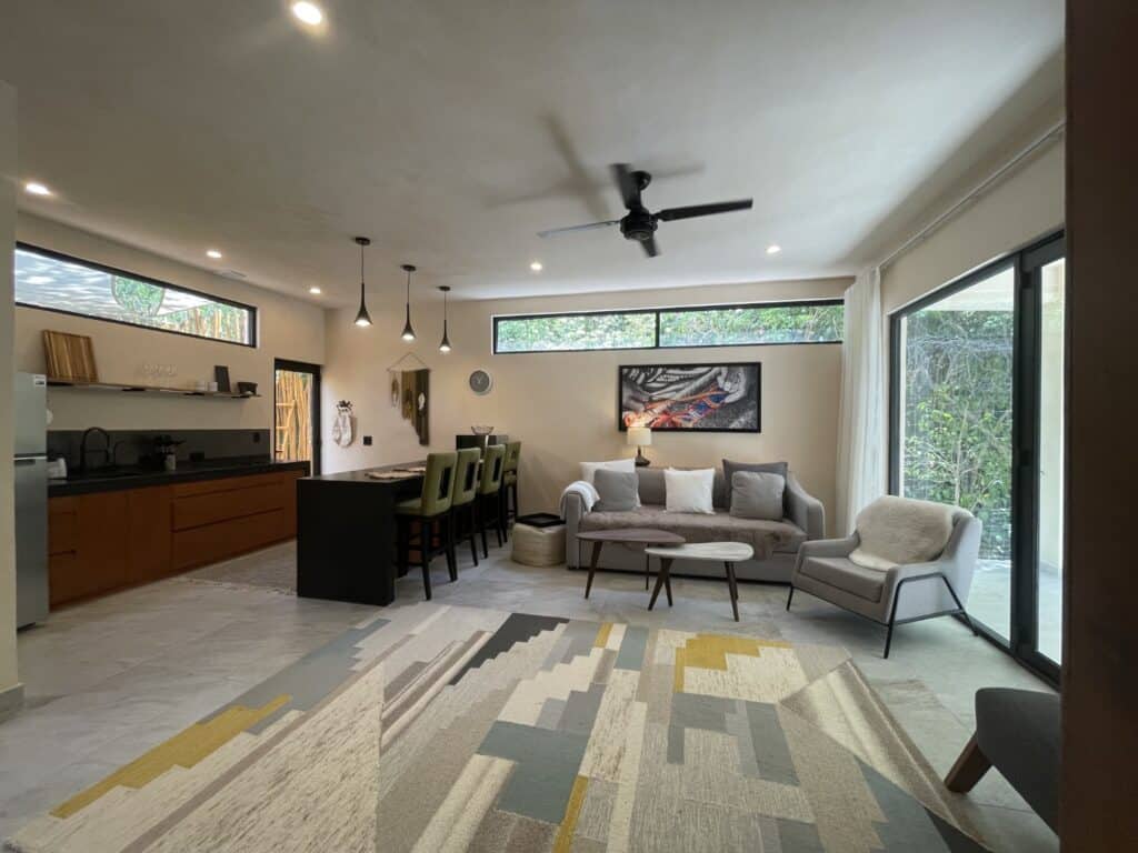 Open-plan living room with large windows, modern furnishings, ceiling fan, and a view of greenery outside at Hacienda MVO in Akumal.