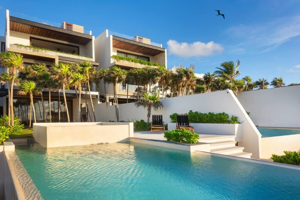 Contemporary beachfront house with a white exterior, a swimming pool, palm trees, and a clear blue sky at Casa Alina in Tulum.
