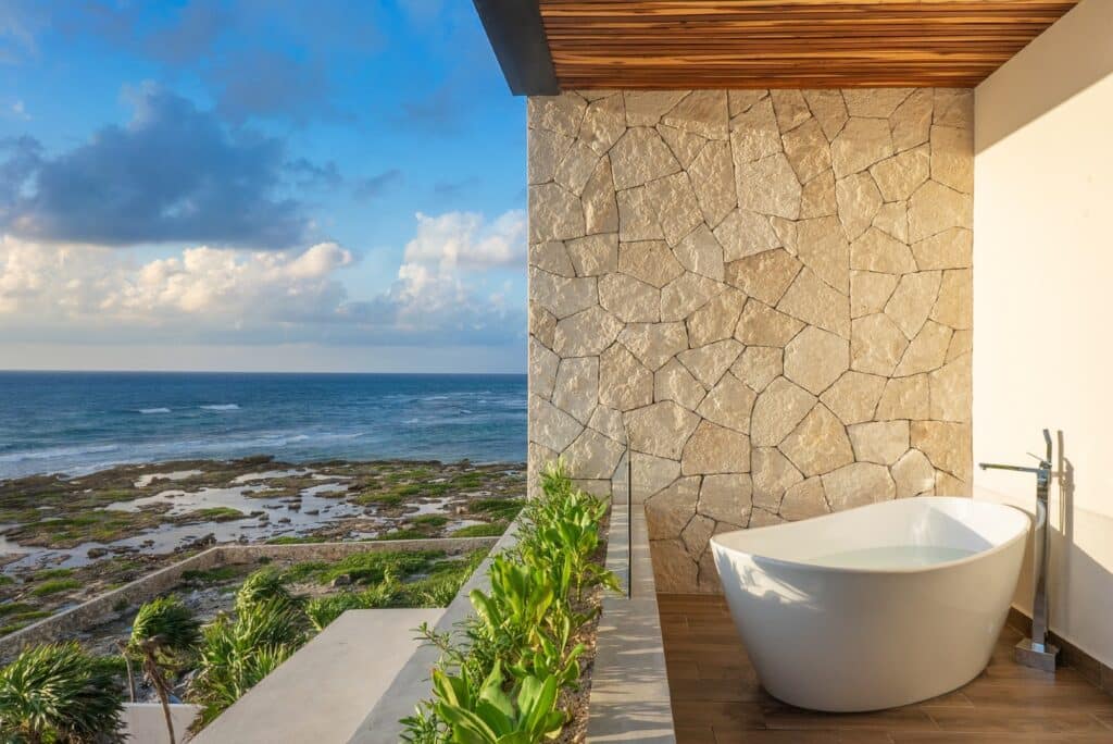 Outdoor bathtub on a balcony with a view of the ocean, rocky shore, and partly cloudy sky during sunset at Casa Alina in Tulum.