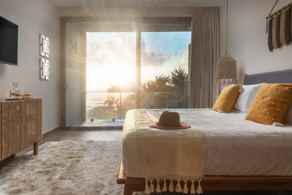 Bedroom with a view of the ocean through glass sliding doors, decorated with mustard-colored pillows and woven accents at Casa Alina in Tulum.