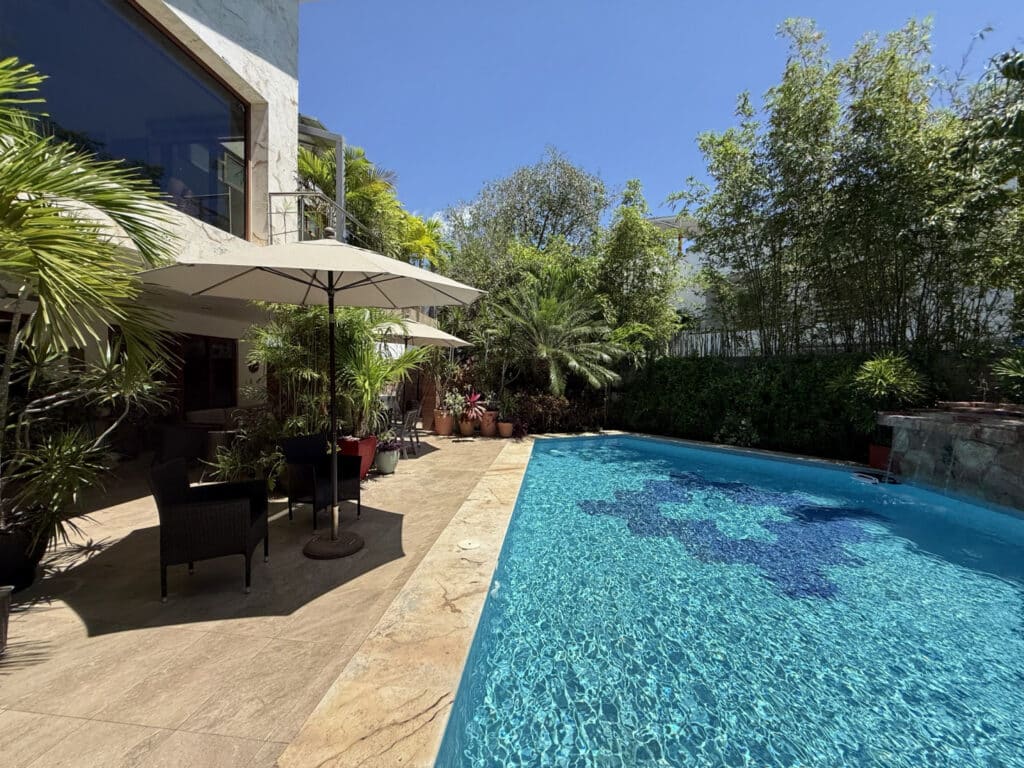Pool area surrounded by lush plants and outdoor furniture, with a modern house and clear blue sky in Playa del Carmen at Casa Noyes.