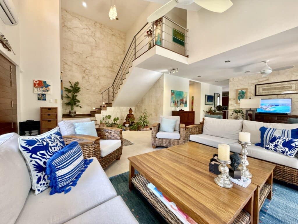 Bright living room with stone walls, wicker furniture, vibrant cushions, potted plants, and a staircase leading upstairs in Playa del Carmen at Casa Noyes.