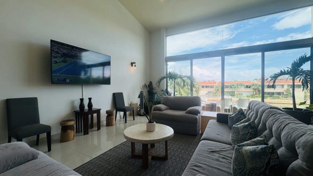 Living room with large windows, comfortable sofas, plants, and TV, overlooking a balcony with outdoor seating and views of neighboring buildings and palm trees.