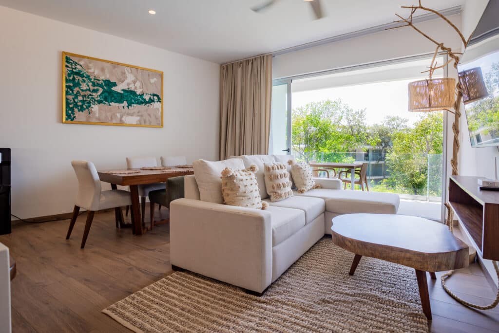 Living room with white sofa, dining area, and glass sliding doors showing outdoor greenery in Playa del Carmen at The Leaf.