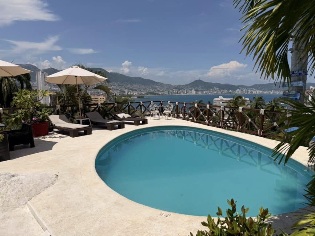 View of a rooftop swimming pool with lounge chairs, umbrellas, and a cityscape with mountains and water in the background in Acapulco.