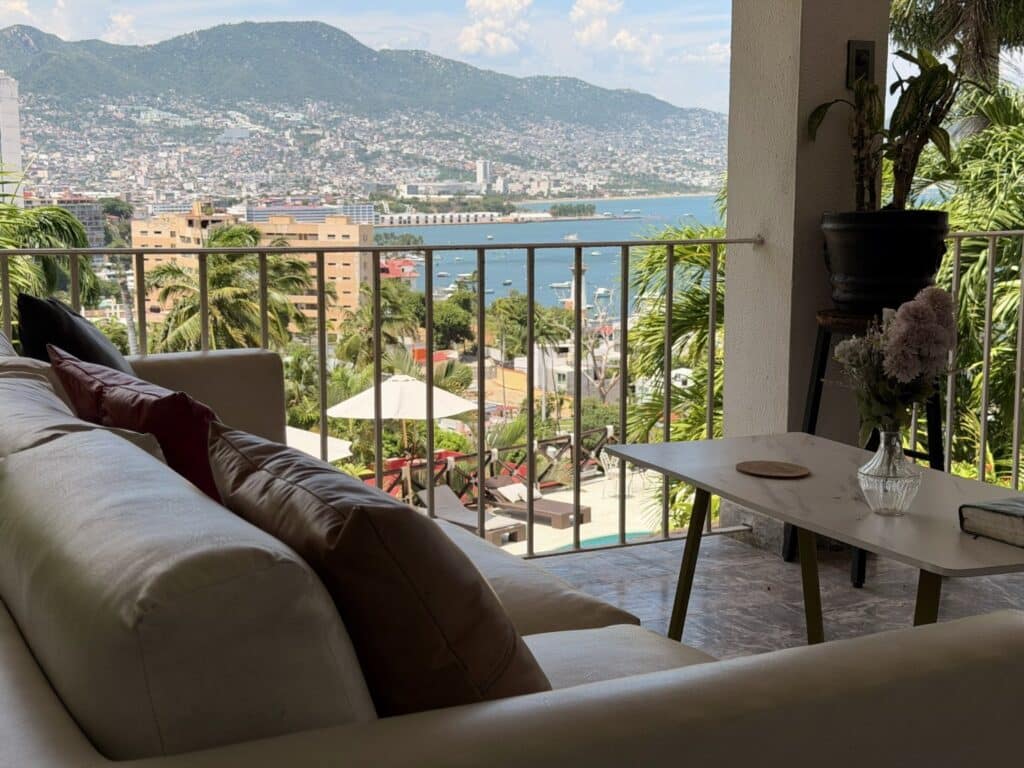 Living room with a balcony view overlooking Acapulco Bay, featuring a cream sofa, a small white table with flowers, and lush greenery.