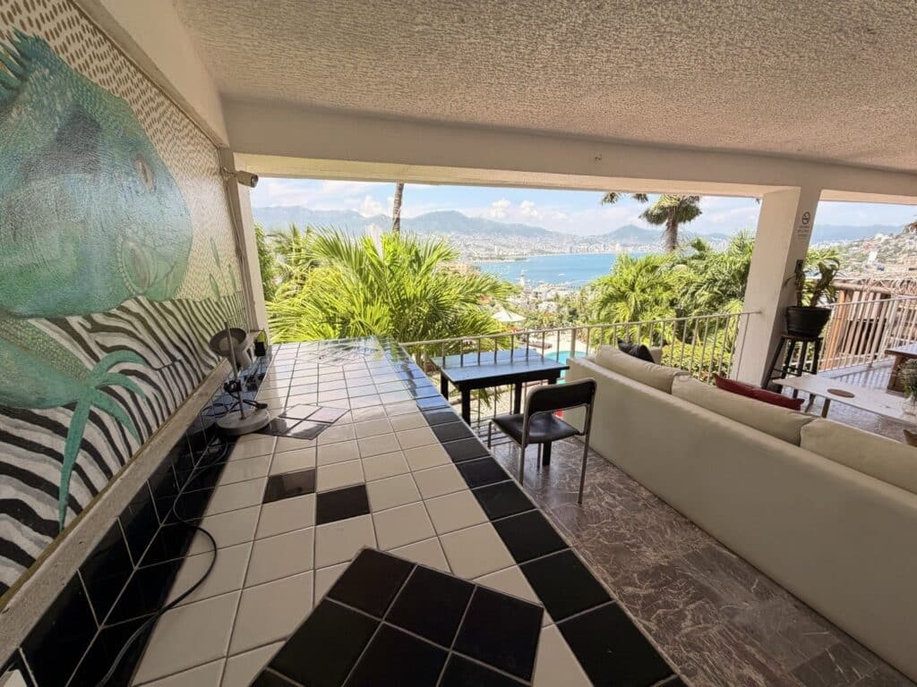 Views of ocean, mountains, and city skyline from a covered outdoor patio in Acapulco, with tropical plants and outdoor furniture.