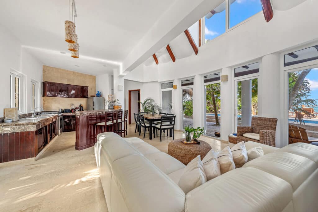 Bright living room with large windows, white walls, exposed wooden beams, and a view of palm tree leaves in Tulum at Casa Sofia.