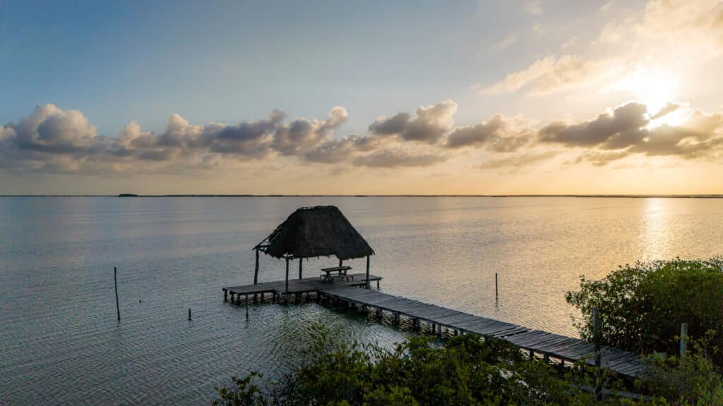 Sunset over tranquil water with a small thatched-roof pier and umbrella, surrounded by lush green foliage in Tulum at Casa Sofia.