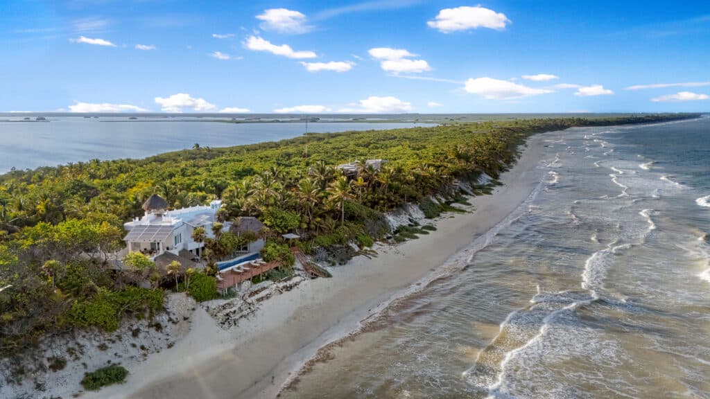 Aerial view of white beachfront houses amid lush greenery along a sandy coastline in Tulum, with blue sky and calm ocean waves at Casa Sofia.