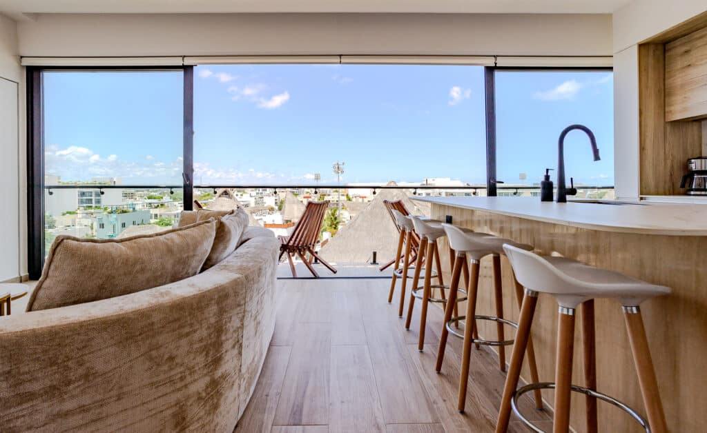 Living room with large glass sliding doors offering a city view, dining area, and a kitchen island with barstools, inside Luna in Playa del Carmen.